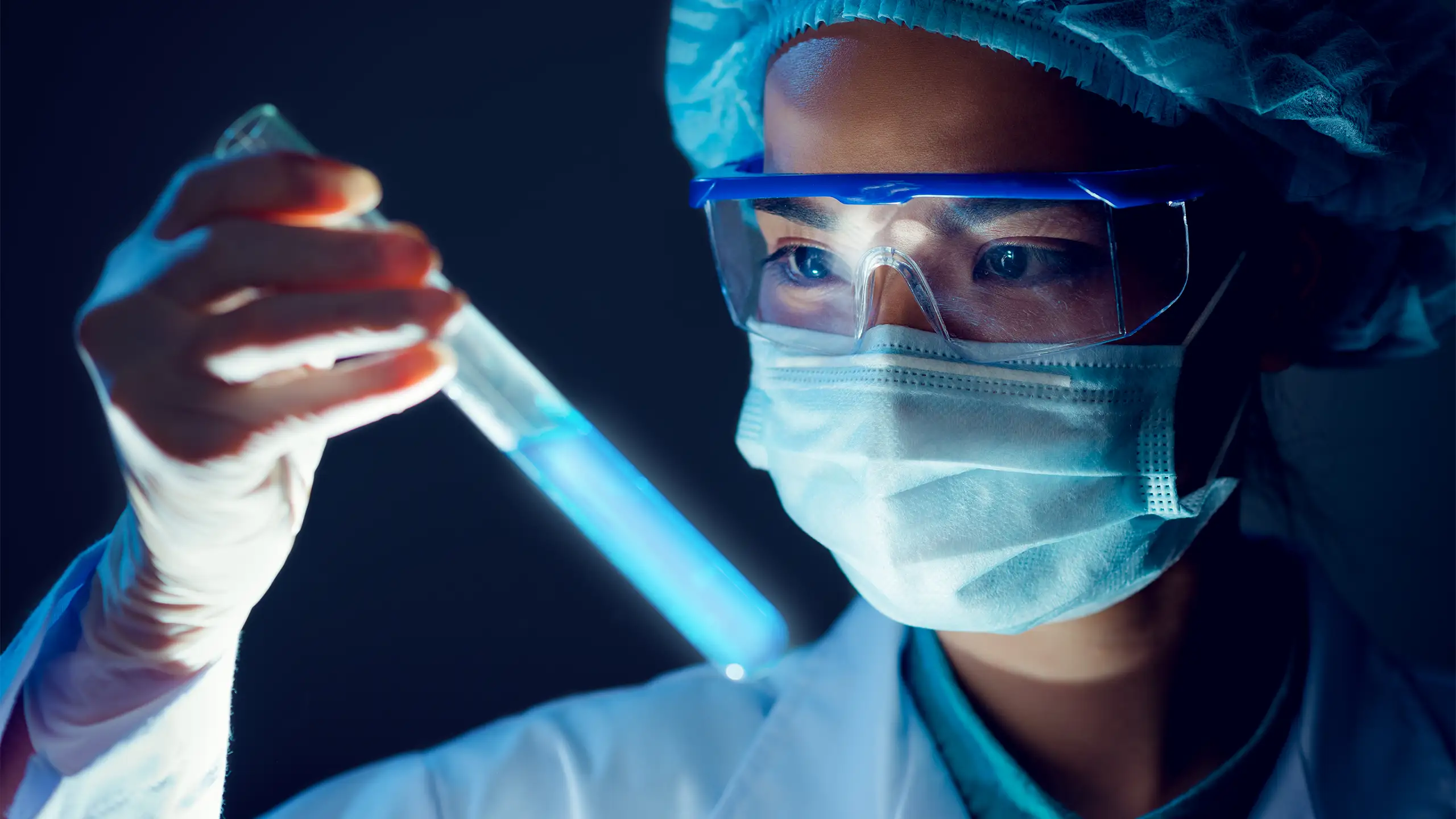 Close-up of a scientist in goggles and mask holding a glowing blue test tube.